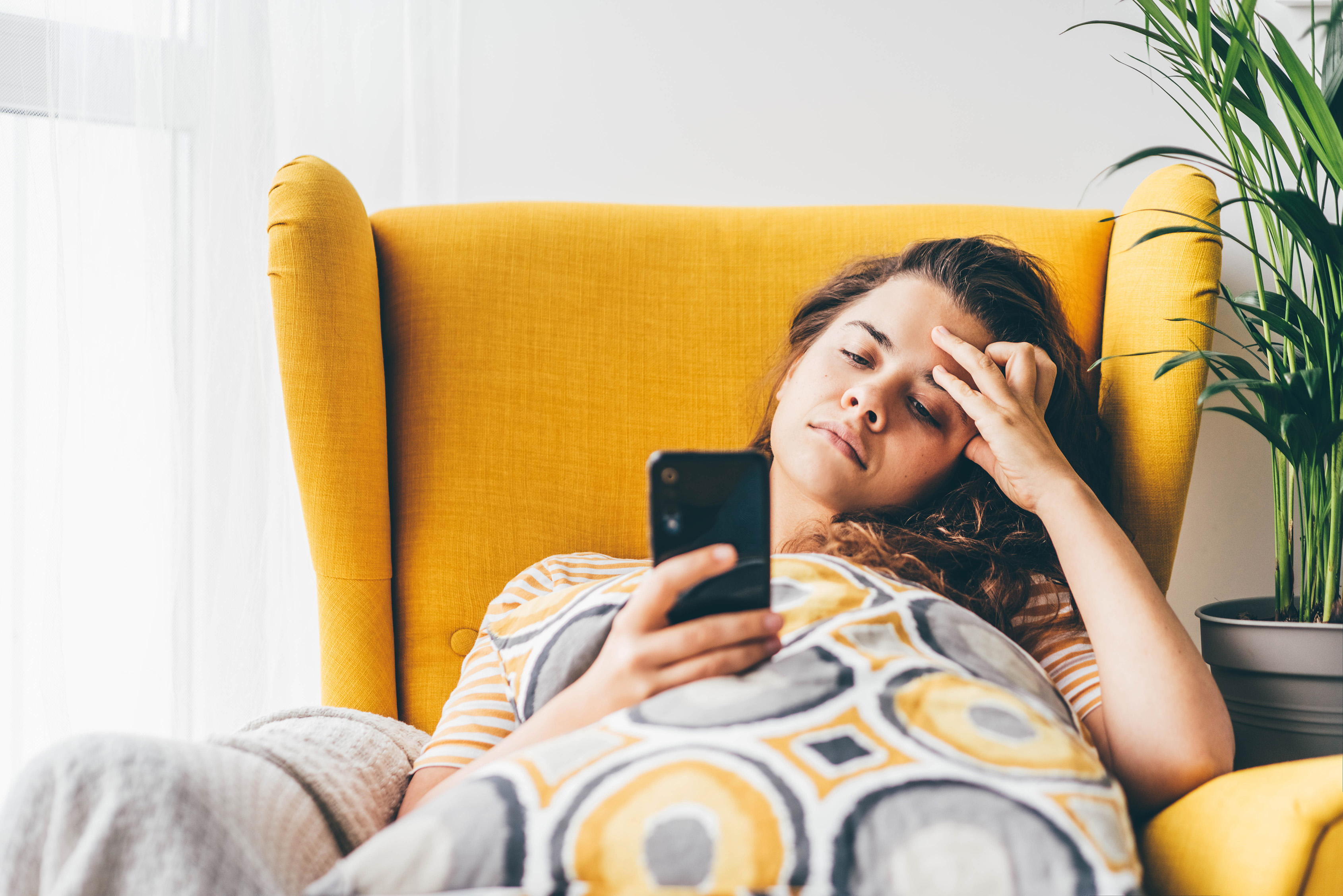 Enttäuschte junge Frau hängt in einem gelben Ohrensessel. Sie ist mit einer Decke mit Kreismuster zugedeckt.